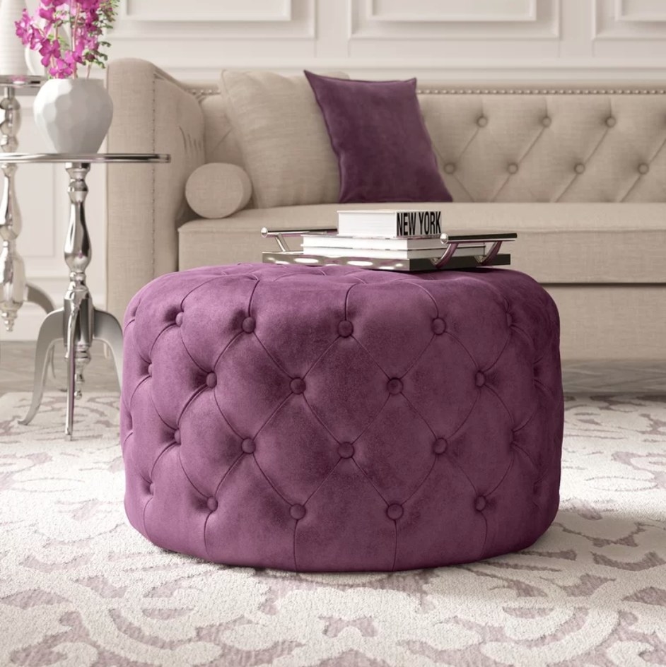 A plum purple, round tufted cocktail ottoman with a serving tray on top filled with books