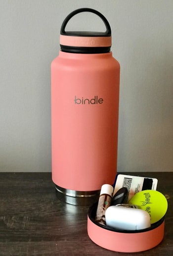 the coral bottle open with belongings such as AirPods, credit card, keys, and lip balm in the bottom compartment of the bottle