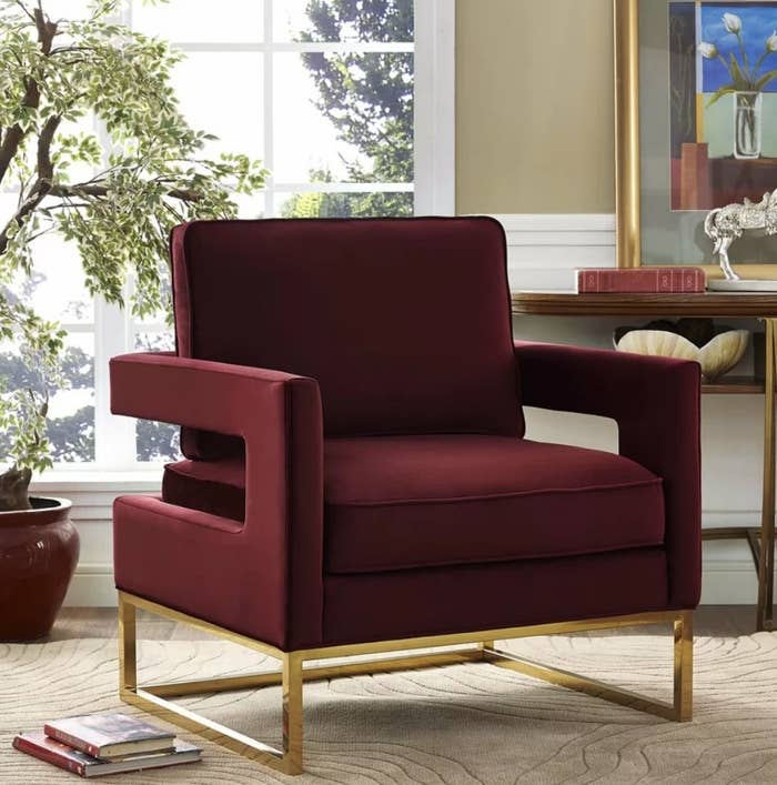 A maroon, velvet armchair with a gold base displayed on a carpet in a living room