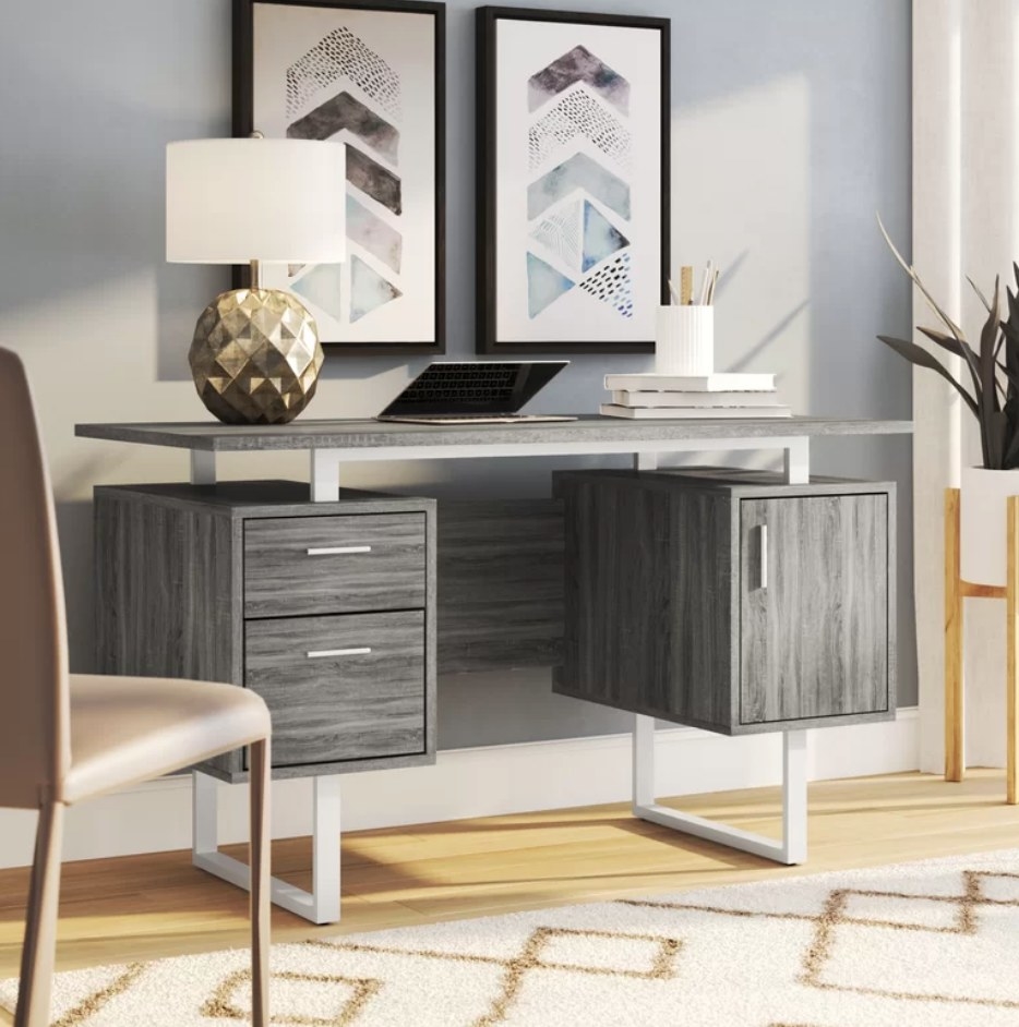 A grey wood desk with two drawers and one cabinet with a laptop and decor atop