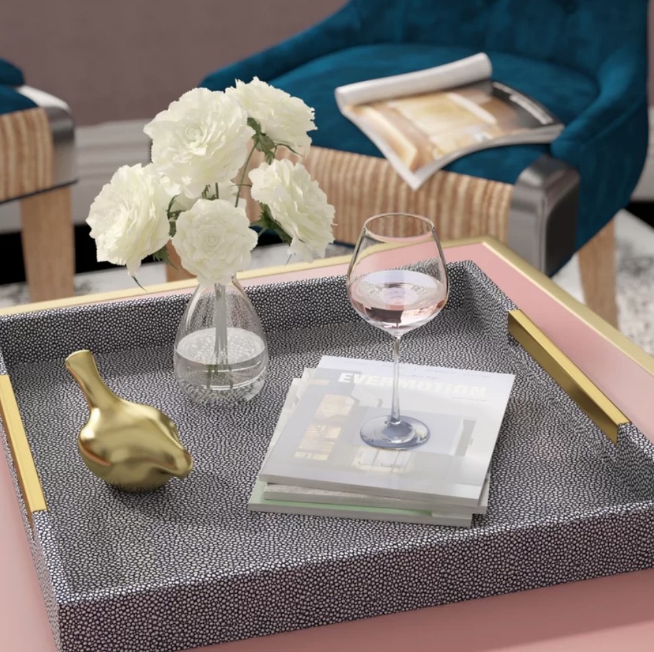 A gray coffee table tray with gold handles atop an ottoman with books, wine glasses, and decor displayed atop