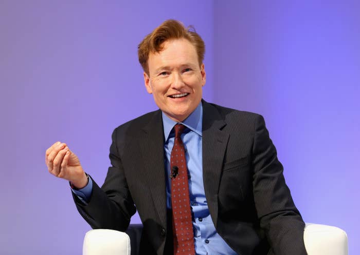 Conan O&#x27;Brien sits in a chair with his hand raised in a black suit, red tie, and blue dress shirt