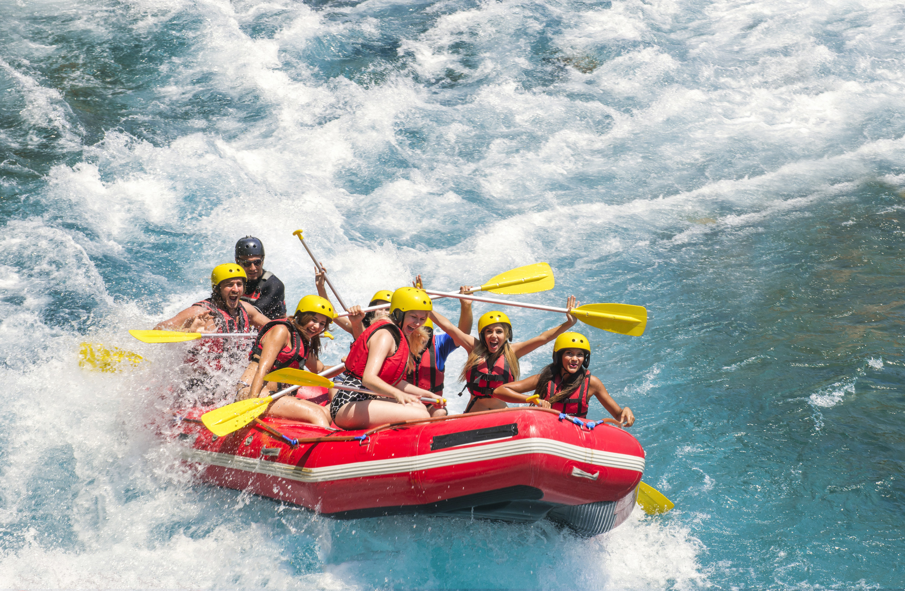 A group of friends white water rafting; they smile as they hurdle down a thunderous part of the river.