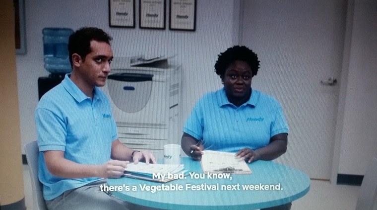 Omar and Stacie are sitting down at a blue table when Stacie mentions a Vegetable Festival.