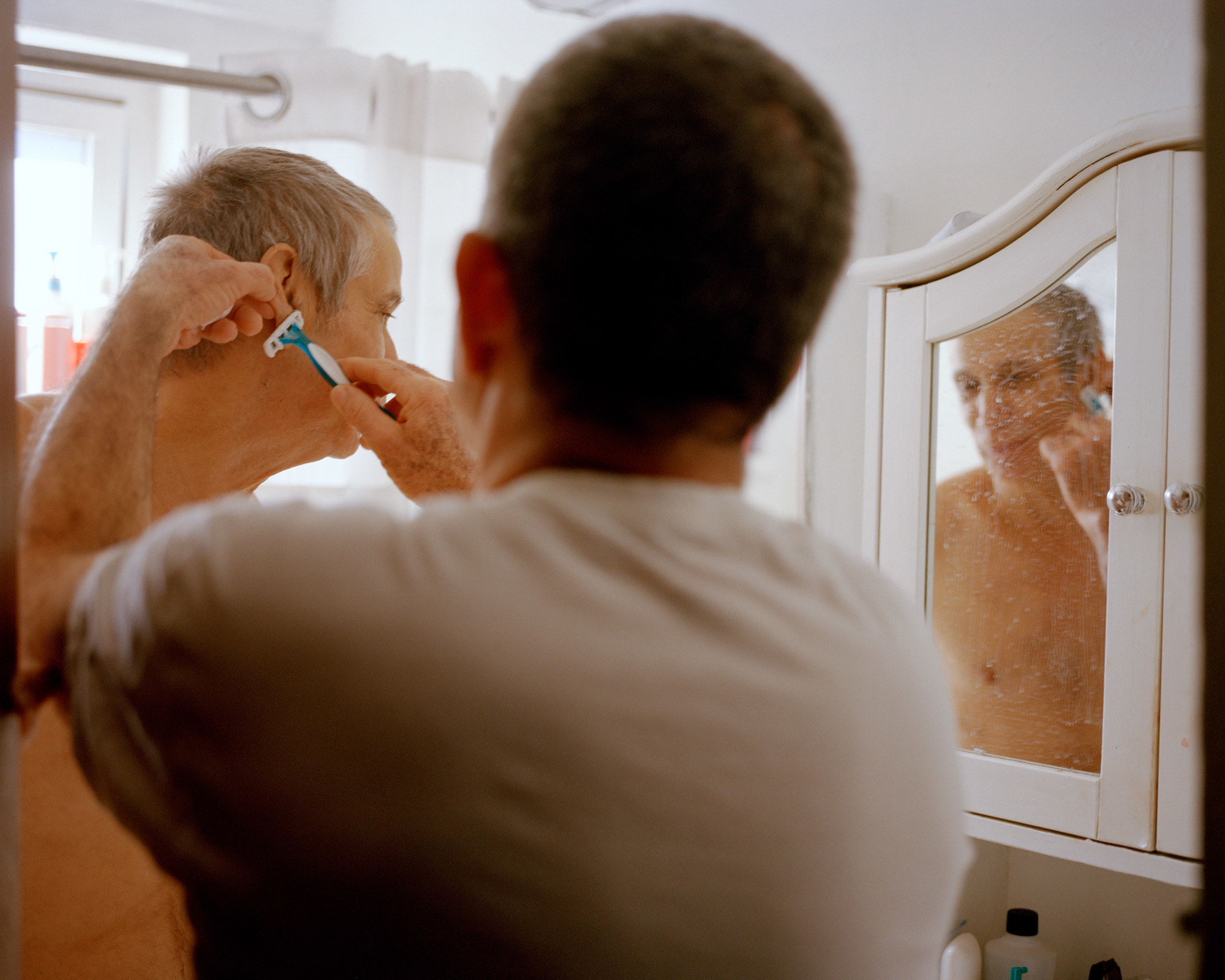 A younger man shaves an older man reflected in the mirror in a bathroom