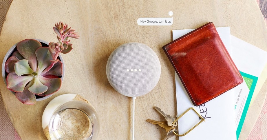Gray nest speaker on table next to wallet, keys, and a houseplant. Speech bubble reads &quot;Hey Google turn it up&quot;