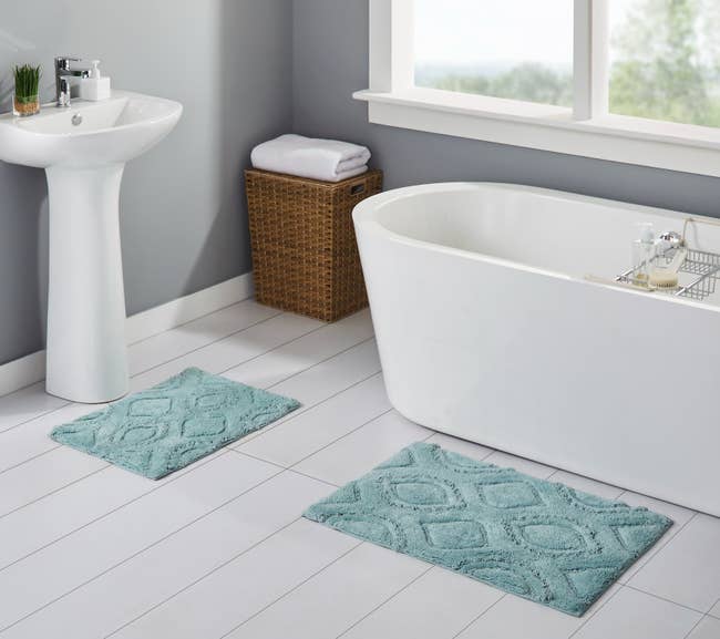 The rug set in a bathroom with the smaller rug underneath a sink and the larger rug just outside a bathtub