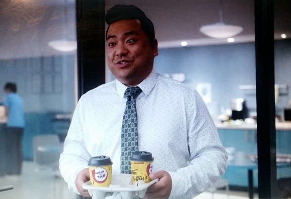 Kimchee, wearing a white dress shirt and tie, carries two Tim Hortons-esque coffees in a cup tray which have red letters on yellow cups