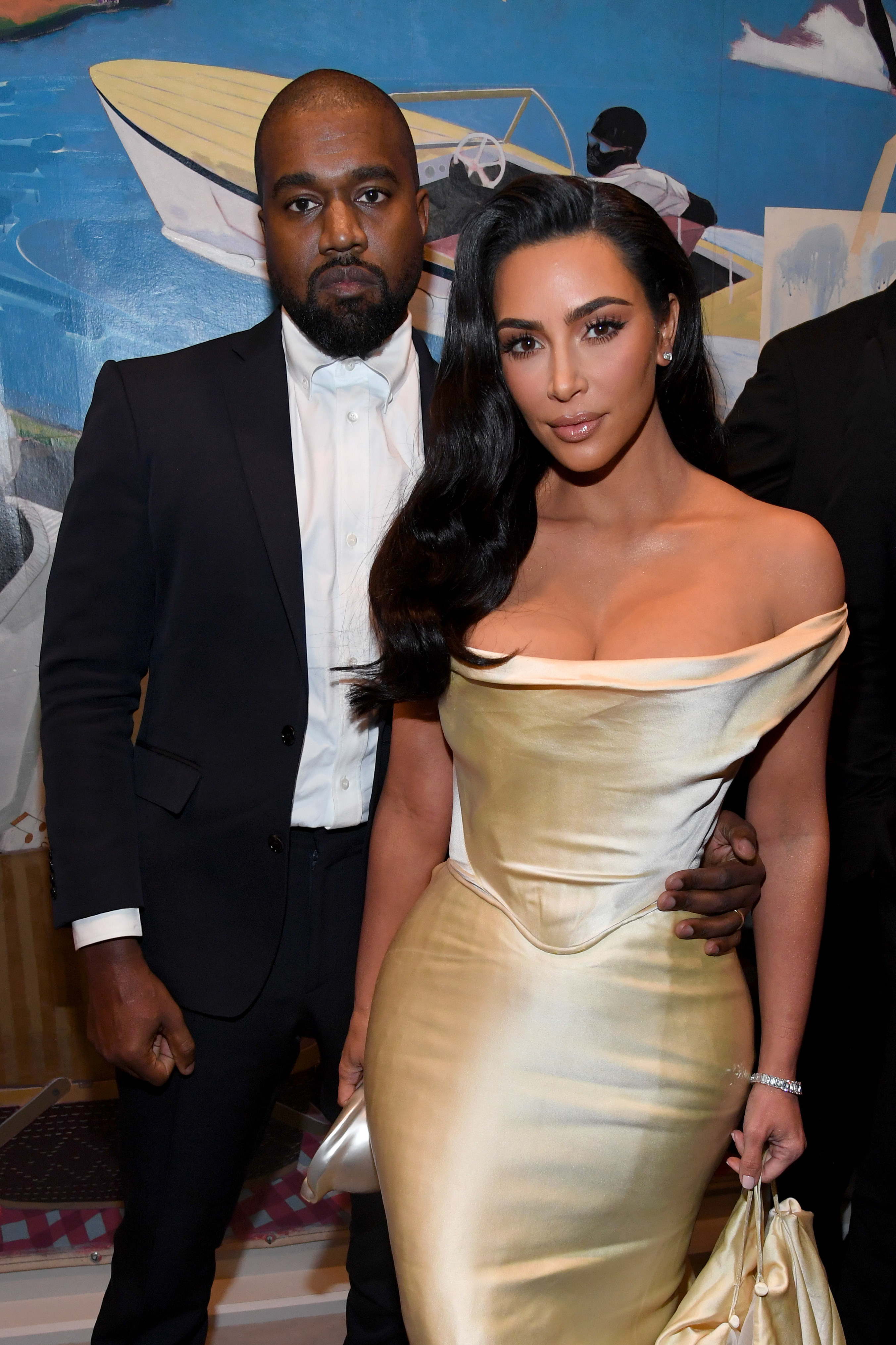 Kanye, in a black suit and white shirt, stands next to Kim, in a gold dress