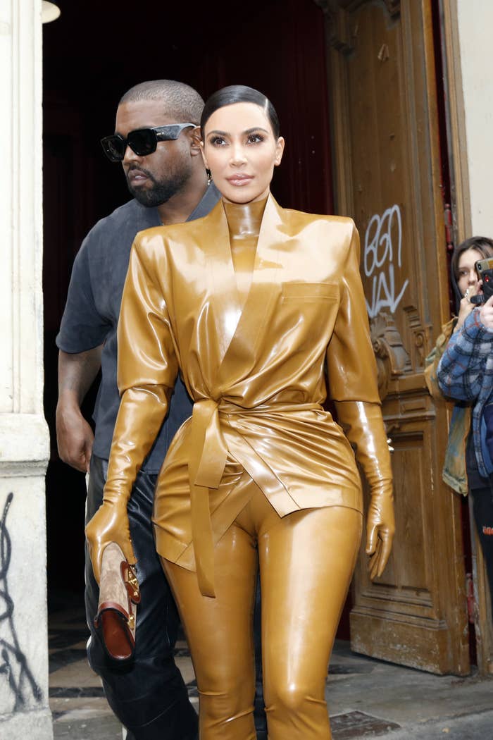 Kim Kardashian, in a yellow leather suit, walks in front of Kanye West, in a black t-shirt and leather pants
