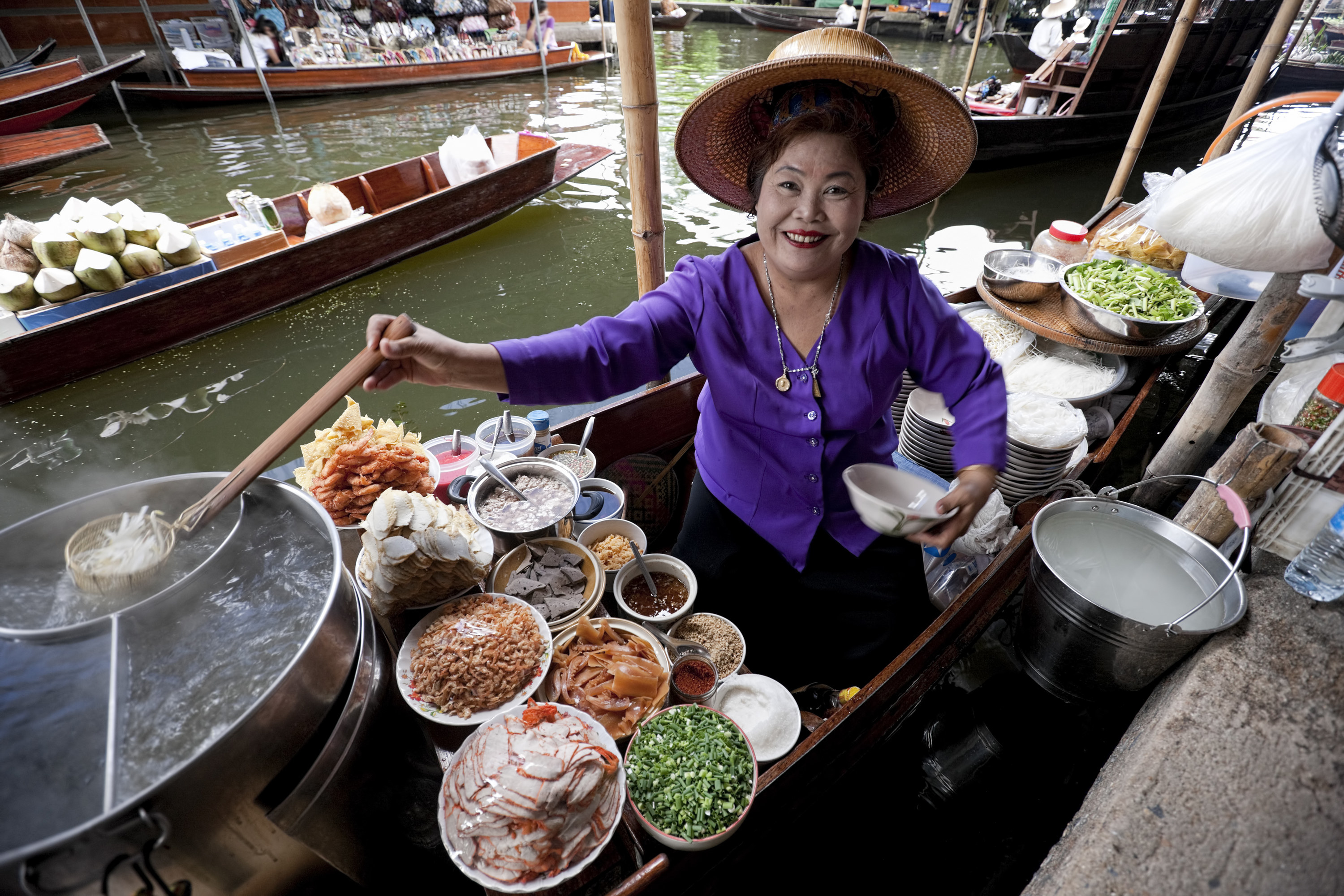 A street foot-vending woman smiles broadly at the camera. She is surrounded by various ingredients and holds a serving of noodles.