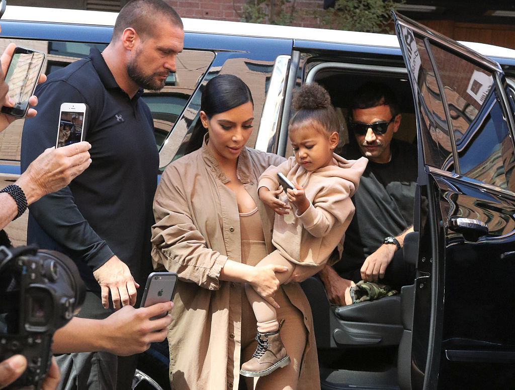 Kim Kardashian is shown getting out of a car, holding her child, as photographers swarm