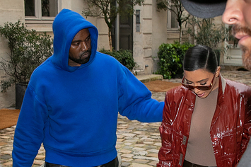 Kanye, in a blue hoodie, puts his arm around Kim, in a red jacket and beige shirt