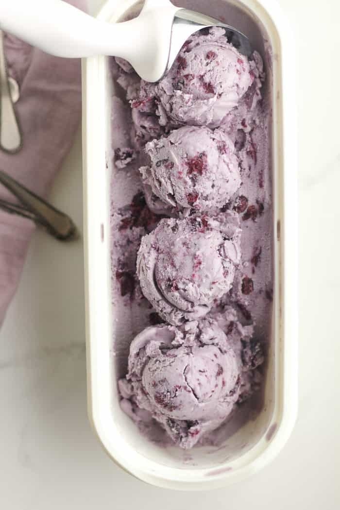 Four scoops of blueberry ice cream in a white dish with a scooper.
