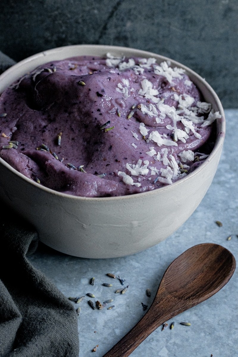 Lavender nice cream in a white bowl next to a wooden spoon.
