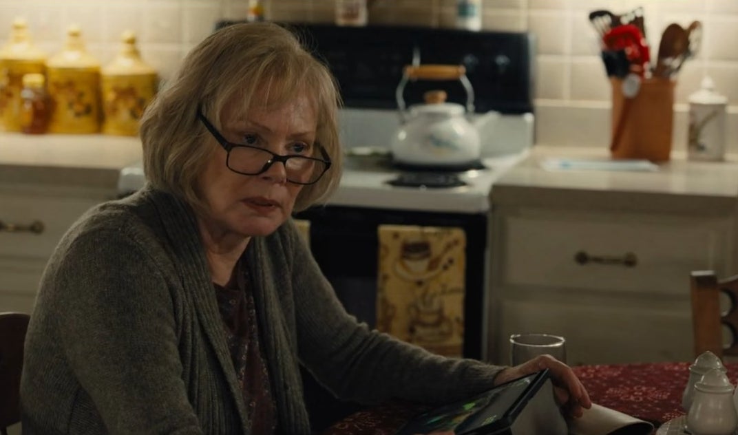 An elderly woman (Jean Smart) wearing glasses, sitting in a kitchen, and staring at something offscreen with a sullen look