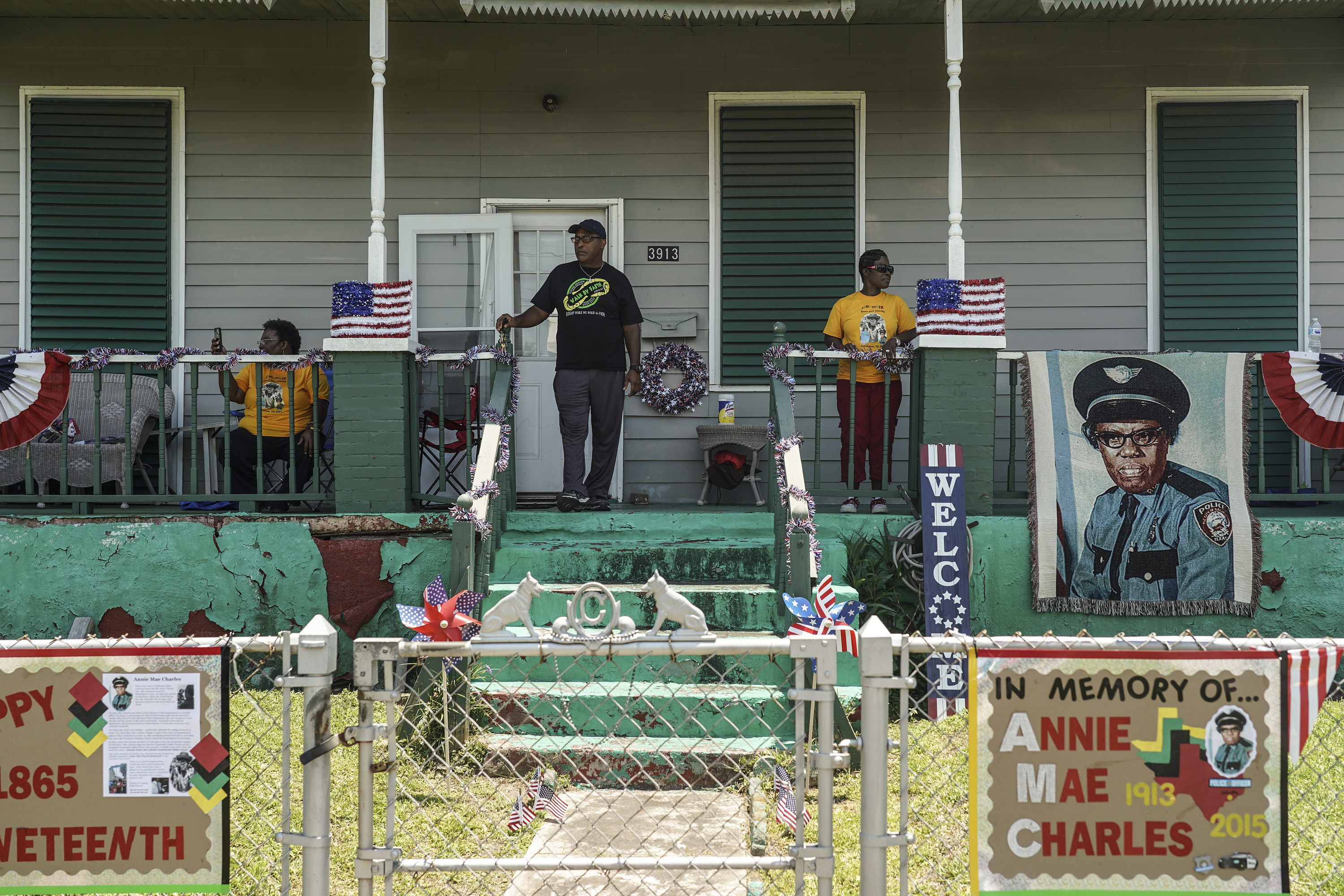 Several people watch from the porch of a house with American flags displayed and commemorative signs. 