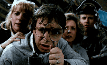 rick moranis and three other adults look closely at the ground, rick moranis has a magnifying glass