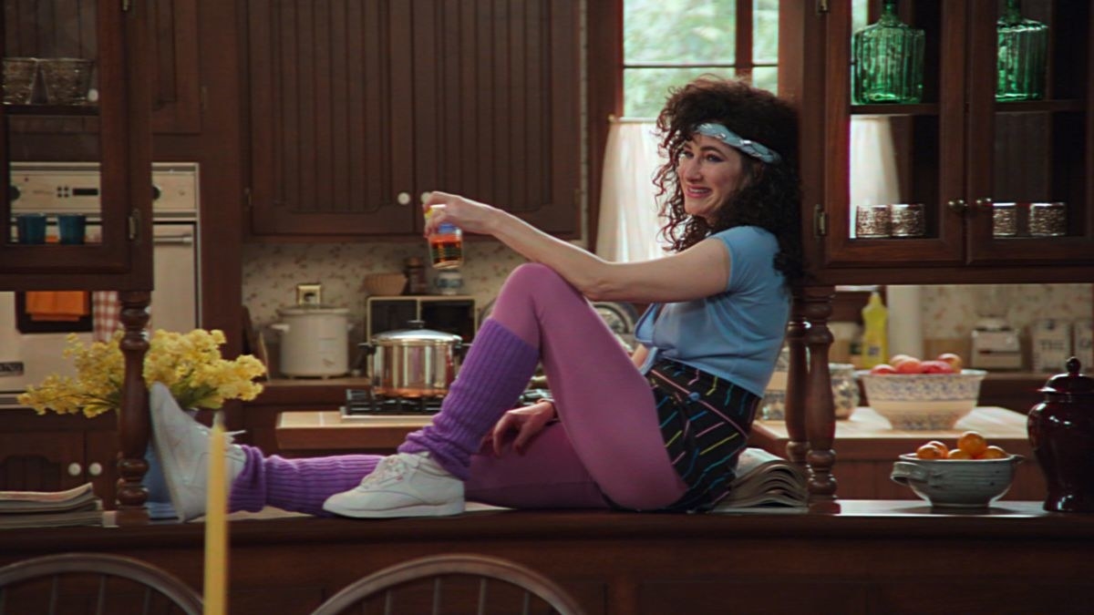 Agatha Harkness (Kathryn Hahn), dressed in &#x27;80s work out attire, sits on a wooden kitchen countertop