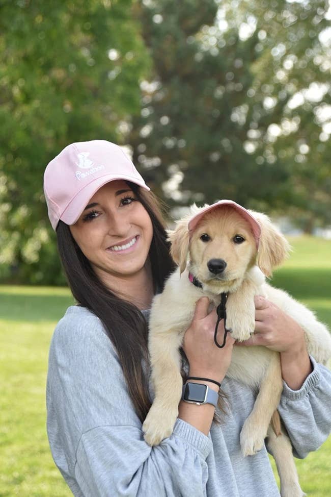A reviewer holding up a puppy, both of them wearing matching pink baseball hats