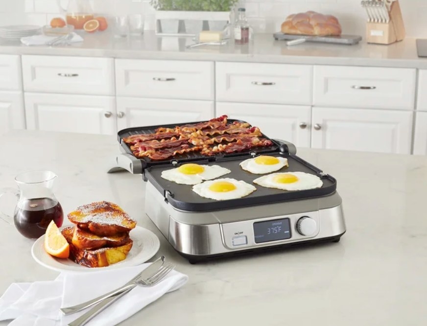 A silver, non-stick, Cuisinart griddler on a kitchen counter filled with eggs and bacon