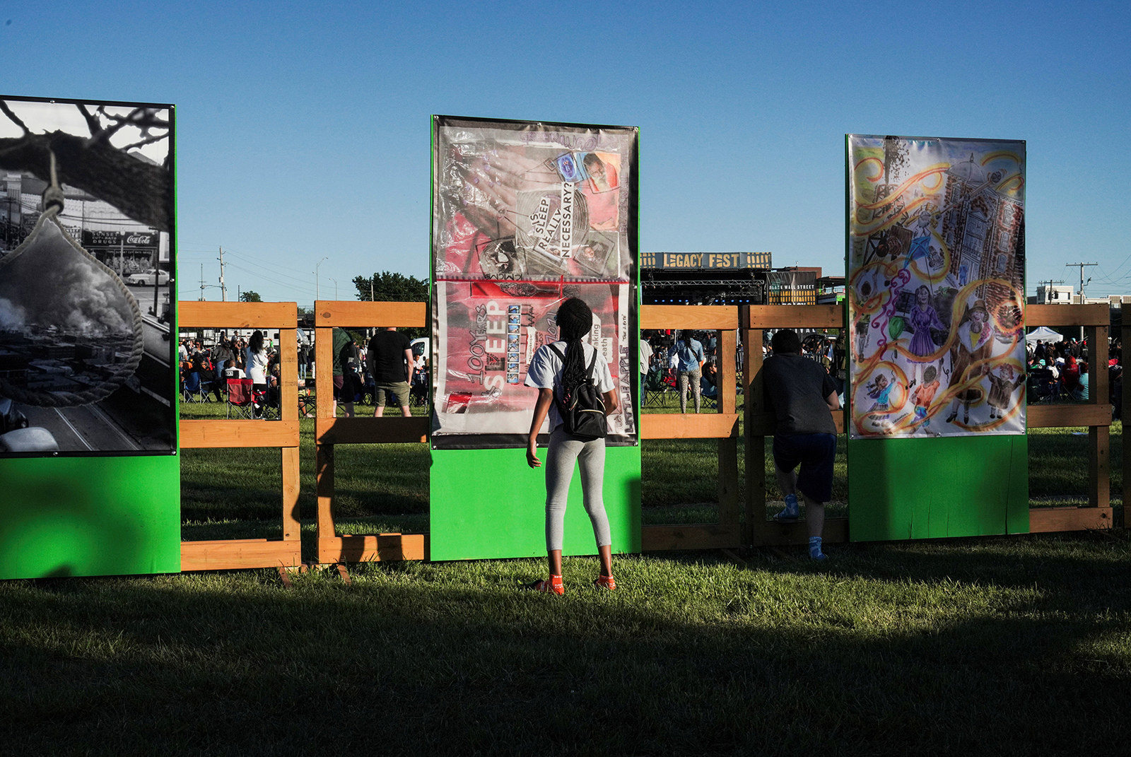 A woman stands behind a fence by three tall vertical artworks that depict the Tulsa massacre through photography, collage, and paint