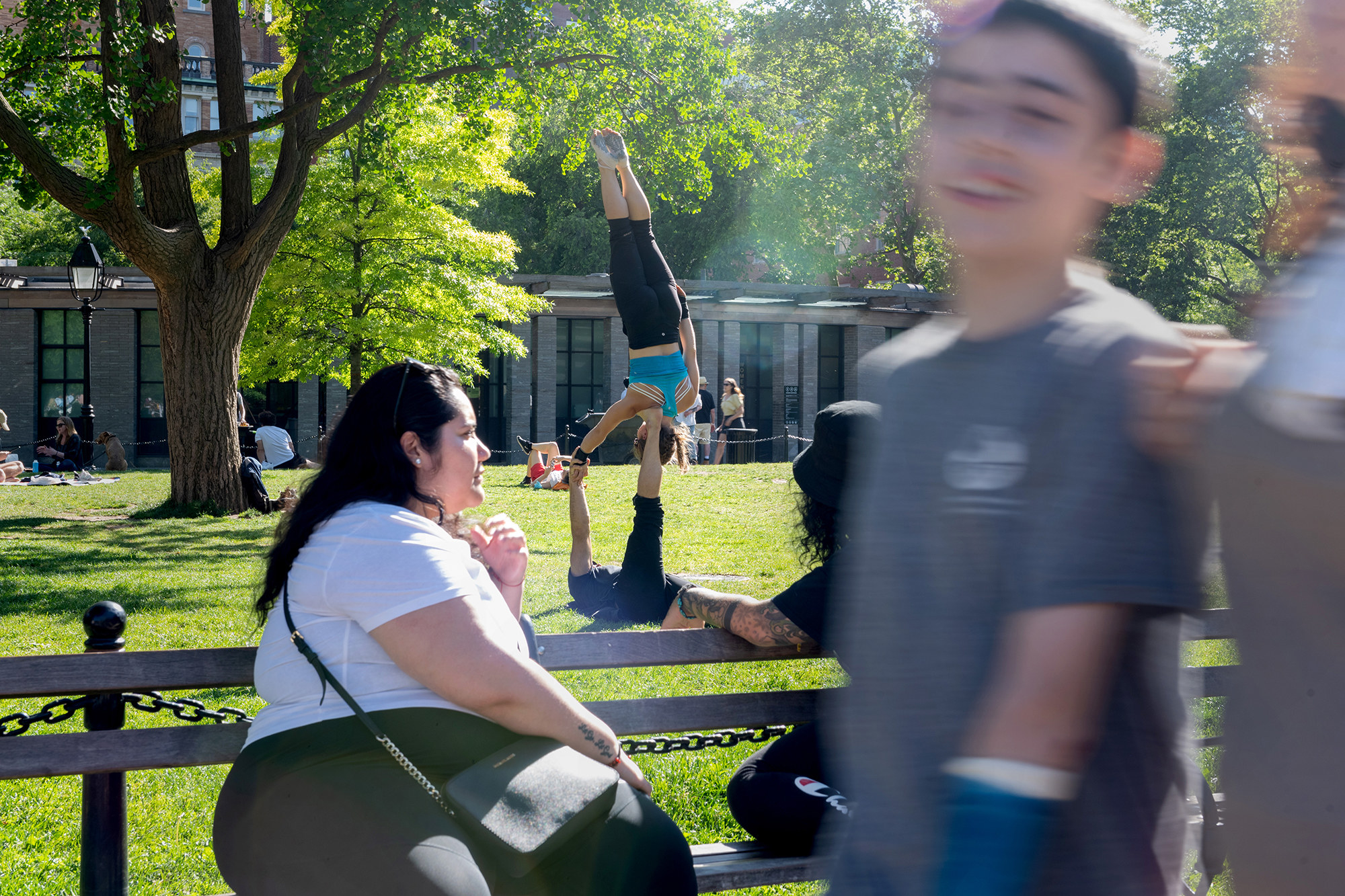 A woman on a bench with her friend watch a couple do acro-yoga, and a boy walks through the forefront of the camera