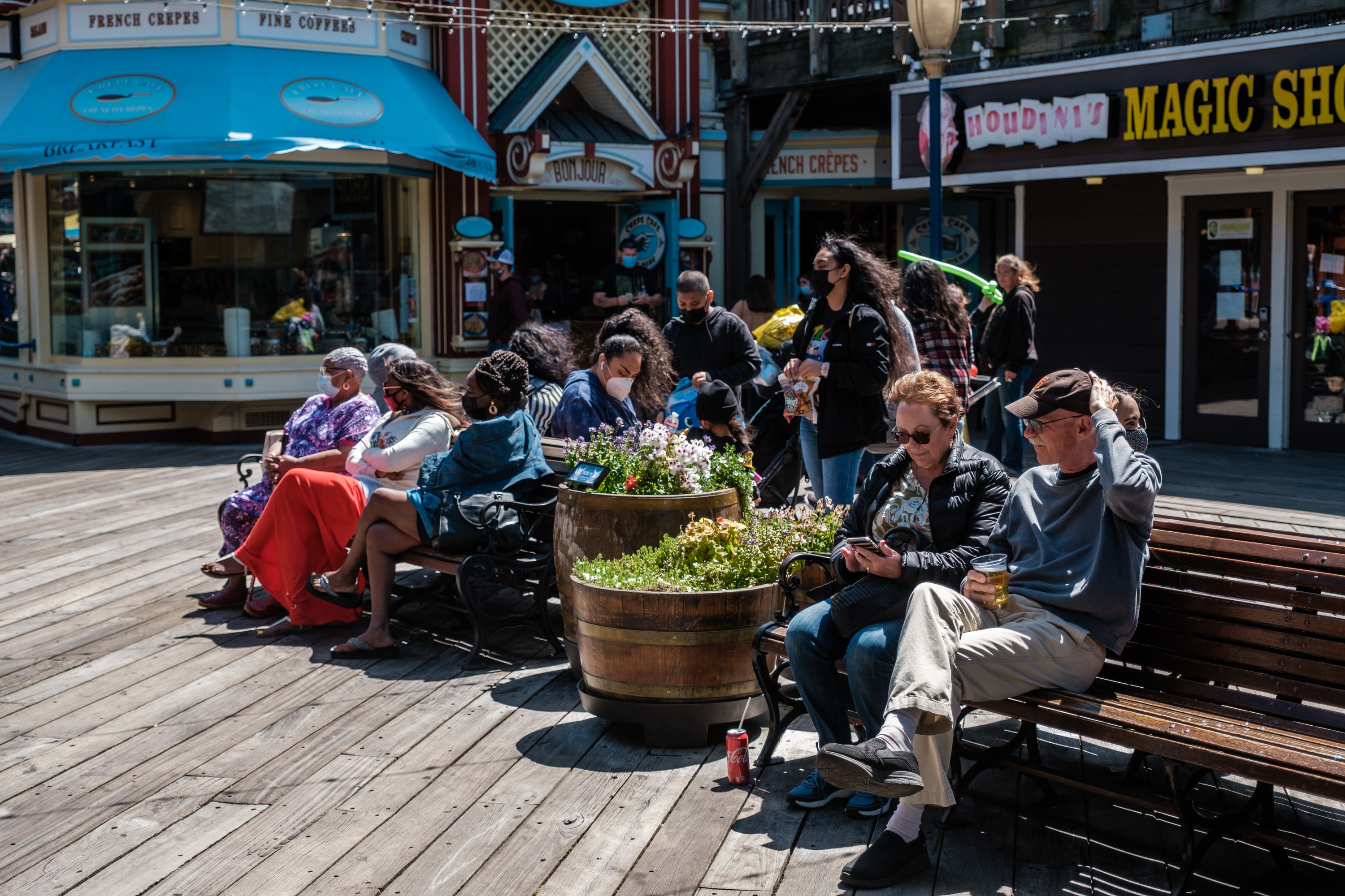 Groups of people sitting on benches in the sunshine on Fisherman's Wharf, some are unmasked