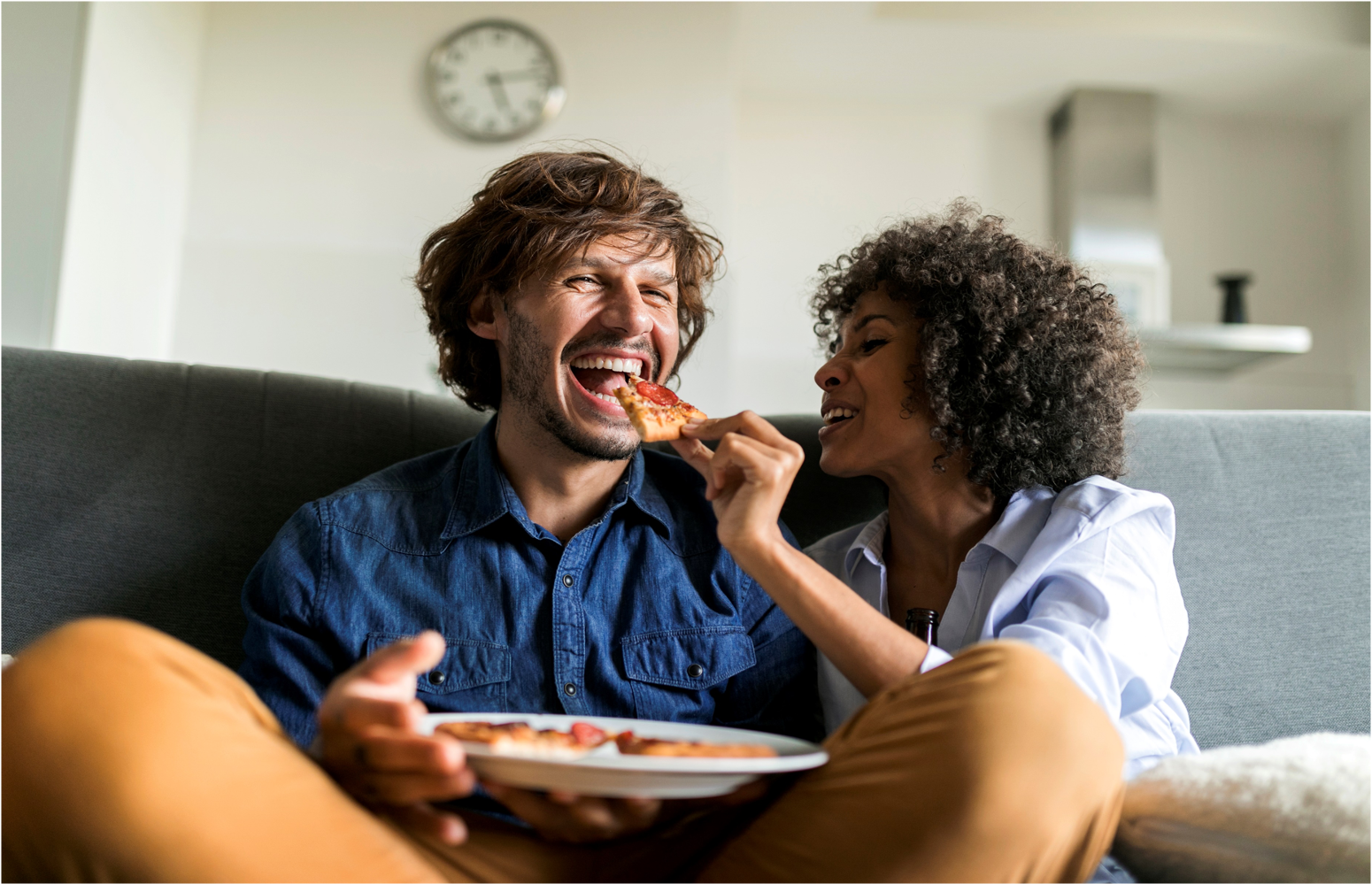 A happy couple eats pizza on their couch.