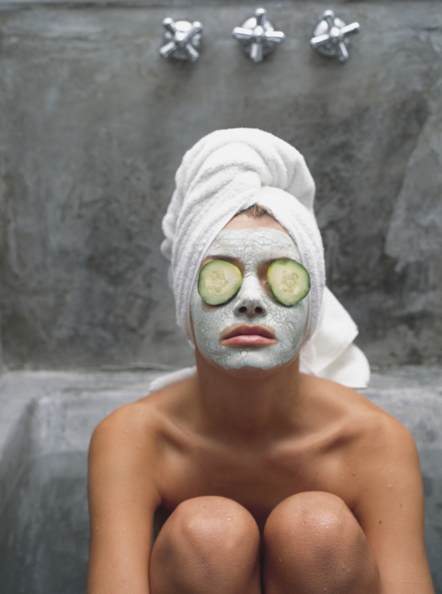 Woman sitting in a spa room with a face mask on and cucumber slices over her eyes, hair wrapped up in a towel