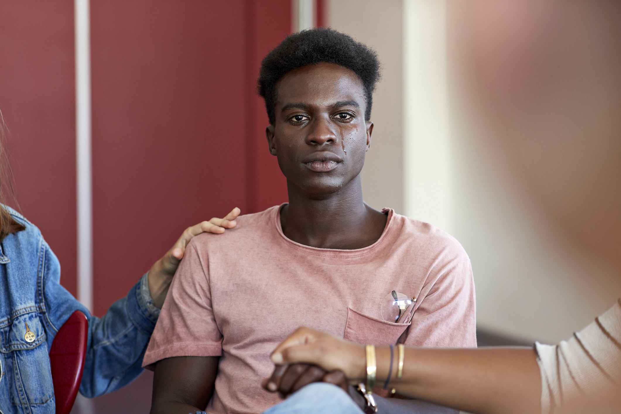 A man in a T-shirt and jeans sitting in a seat, tears running down his face, with the hands of the two people sitting on either side of him patting his shoulder and holding his hand