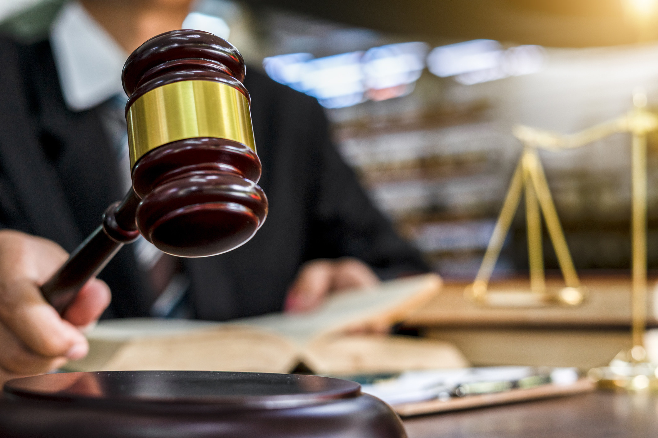 Close-up of a gavel being held by a judge-type holding a book, about to strike a sound block. Golden scales of justice sit on a table in the background