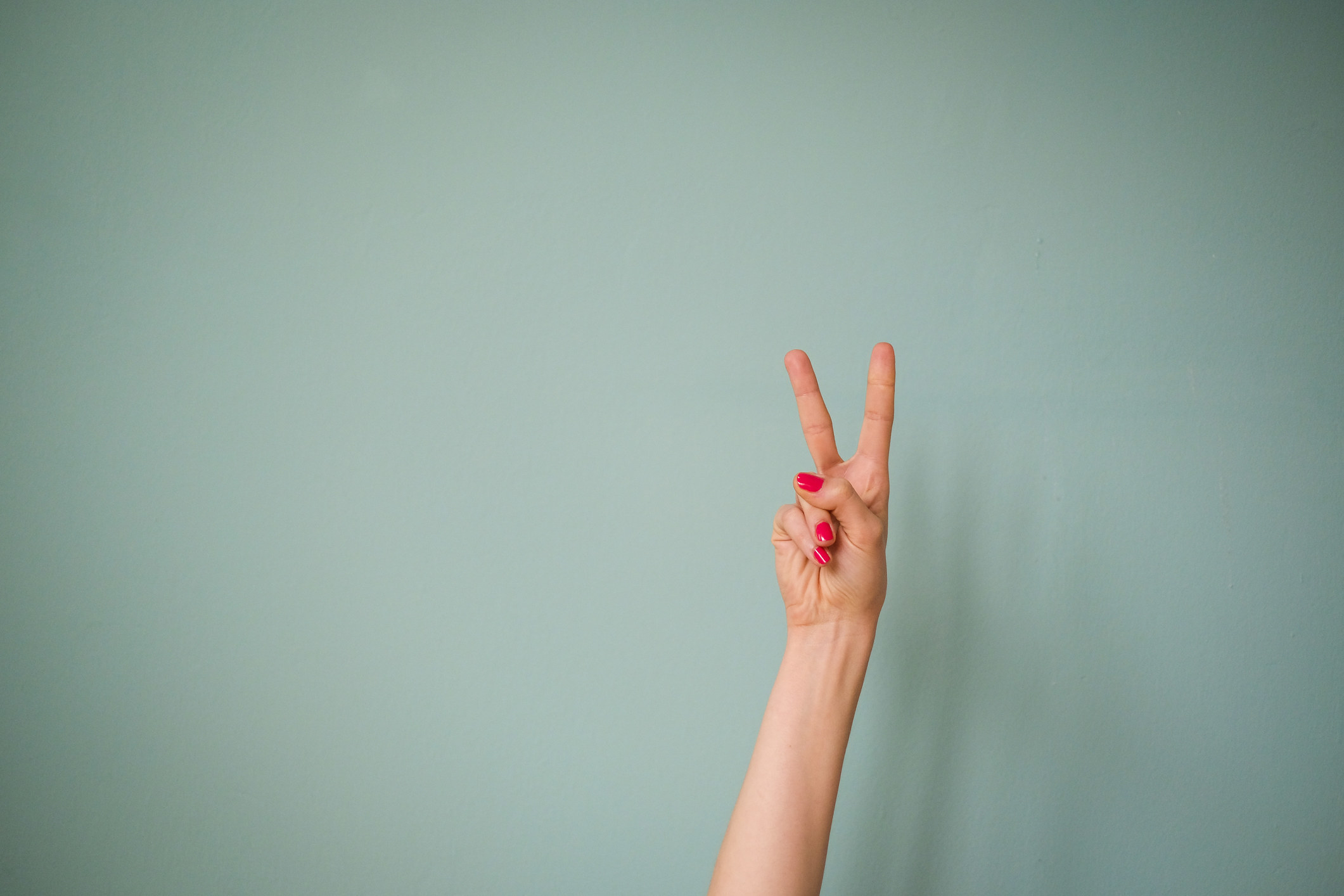 A person holding the peace sign with her nails painted red against a mint colored call