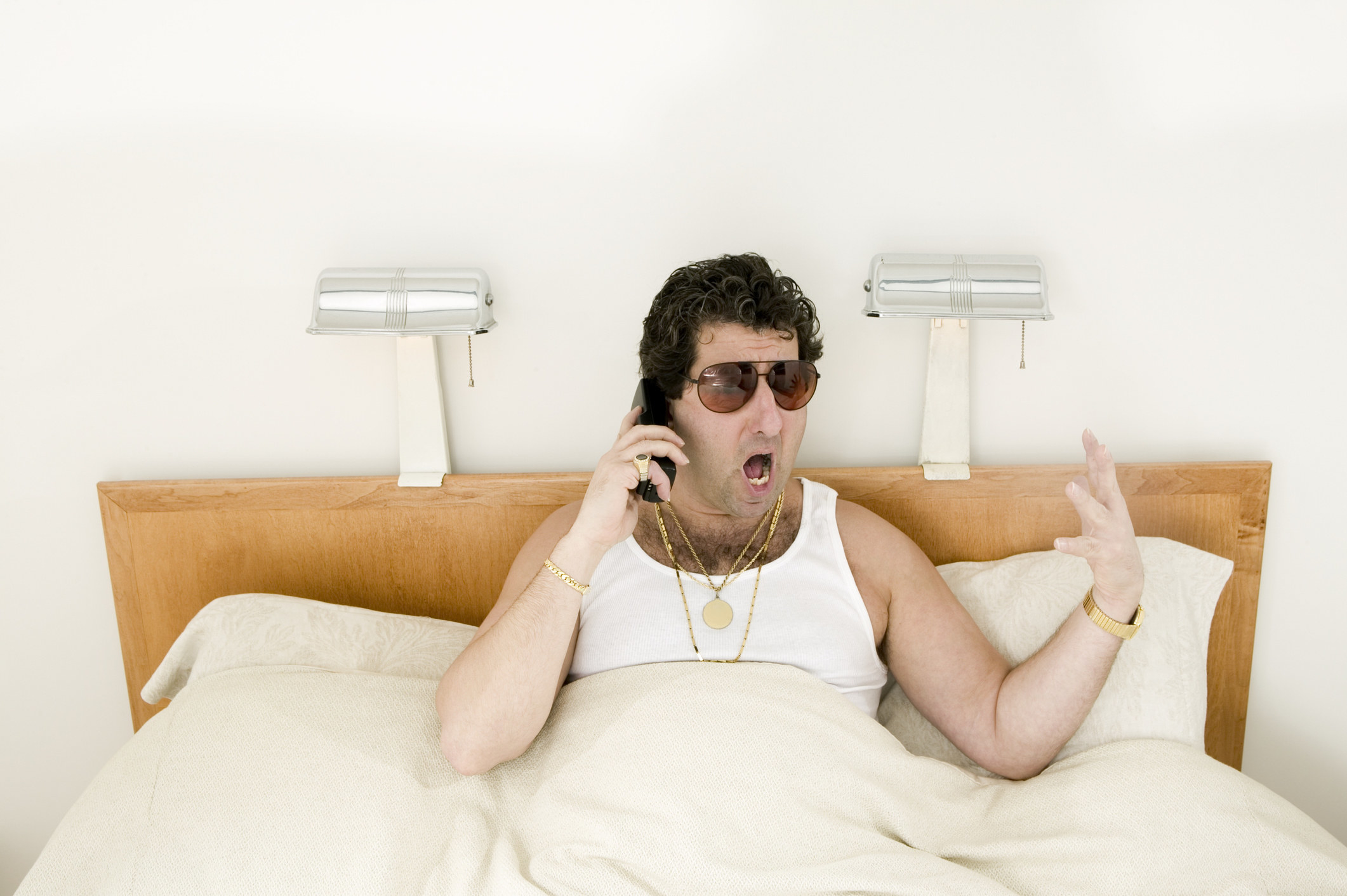 A man in bed wearing a tank top, chains, and sunglasses, yelling into a phone and gesturing with his hand