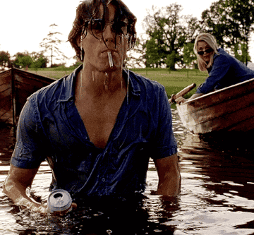 Hugh Grant spitting out water while wading through a lake; he is soaking wet