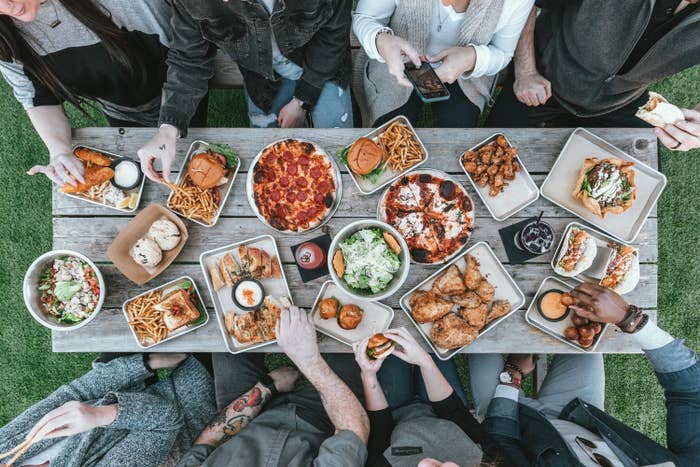 A picnic table full of food, people sitting around the picnic table eating. 