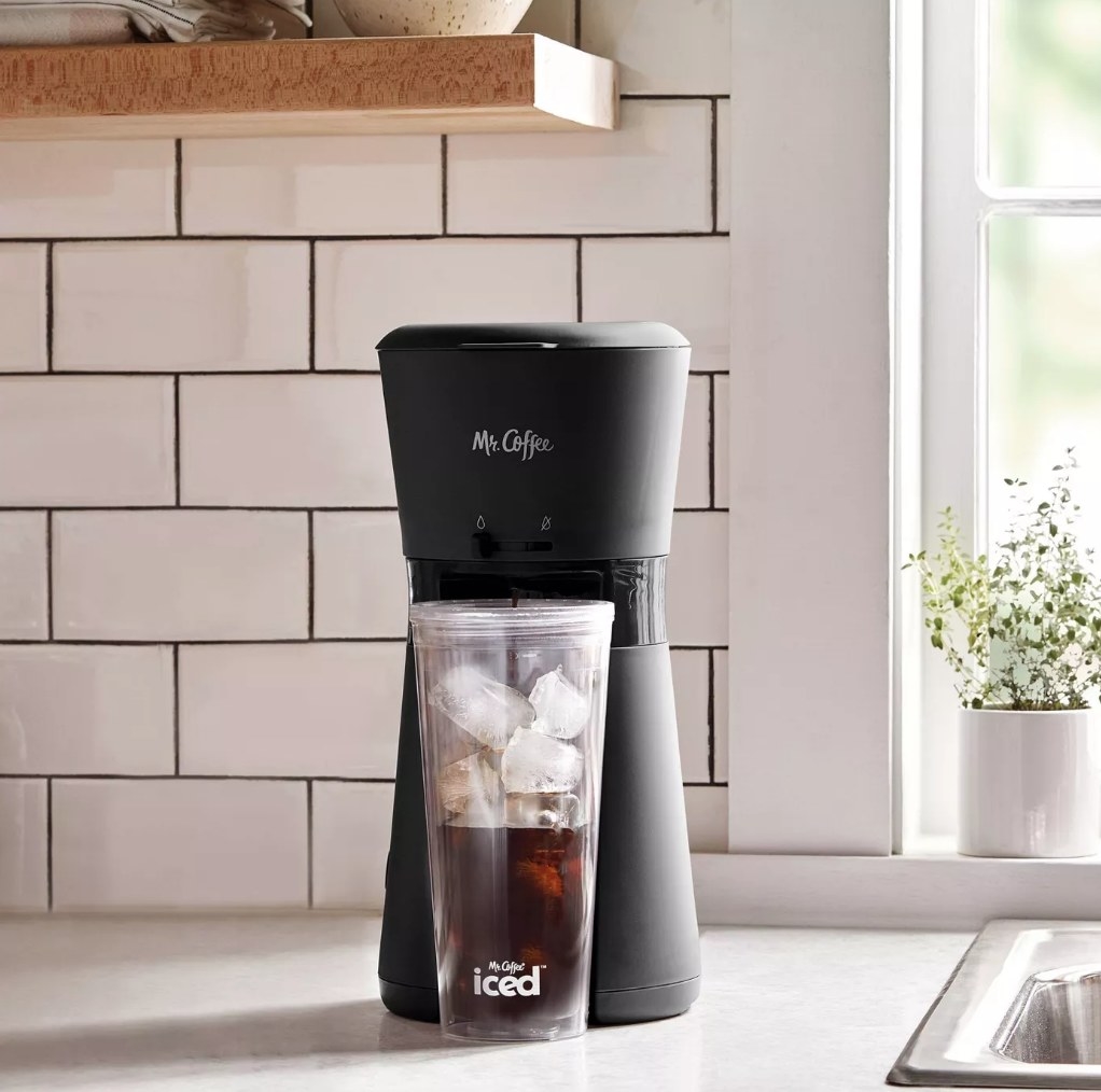 A Mr. Coffee Iced Coffee Maker with Reusable Tumbler and Coffee Filter in black pouring coffee into a tumbler cup