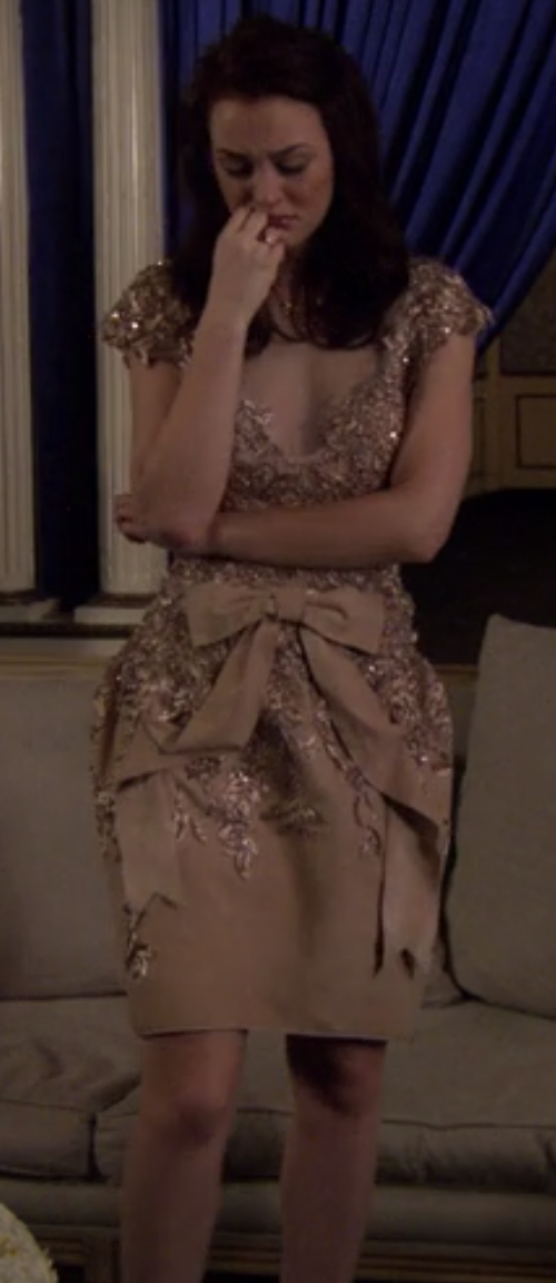 Blair in a short structured dress that goes out at the hips then back in with bows on the skirt, cap sleeves, a sheer overlay over the deep v neck, and sequins going all down the front