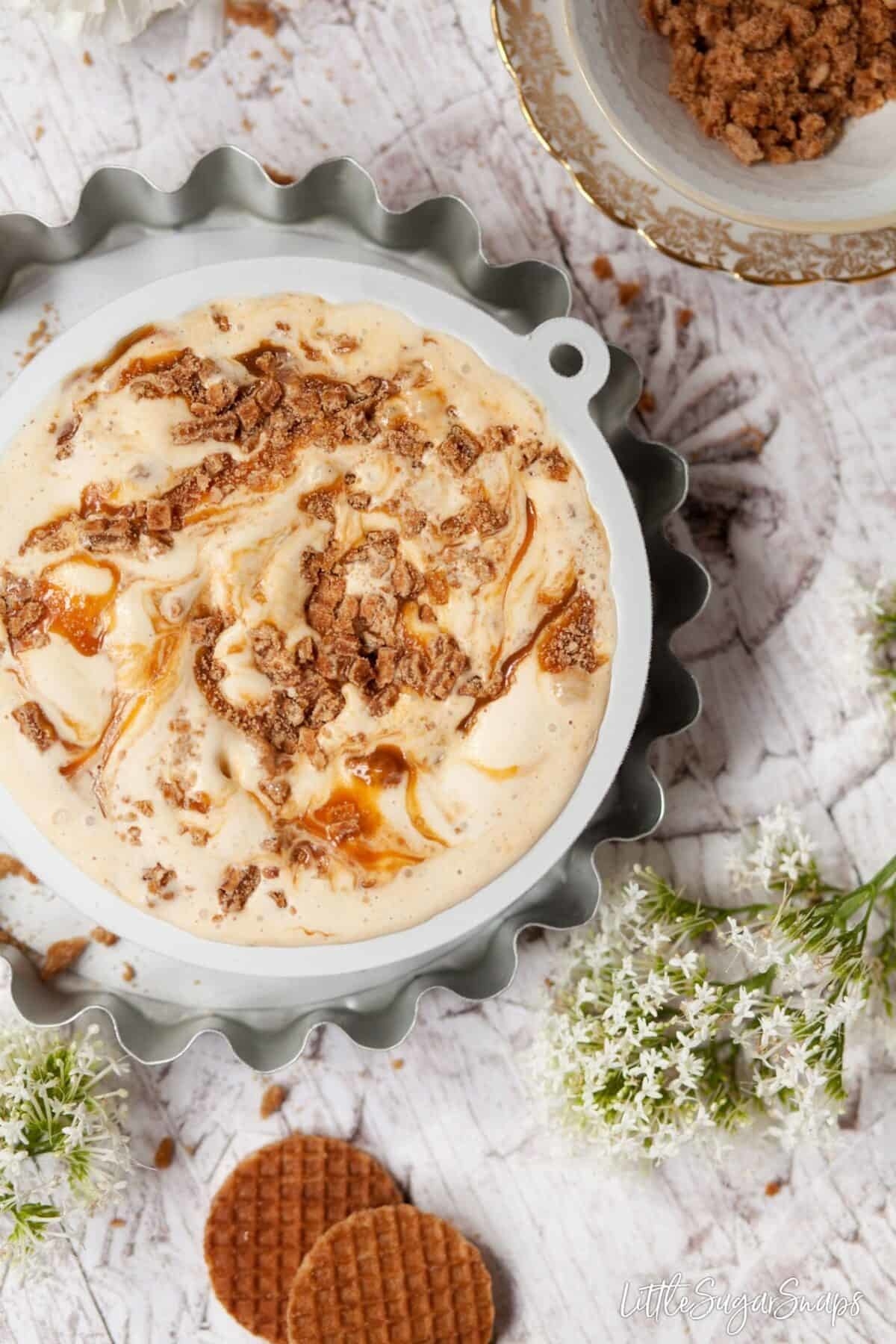 Ice Cream with swirls of caramel and cookie crumbles in a dish next to flowers and cookies.