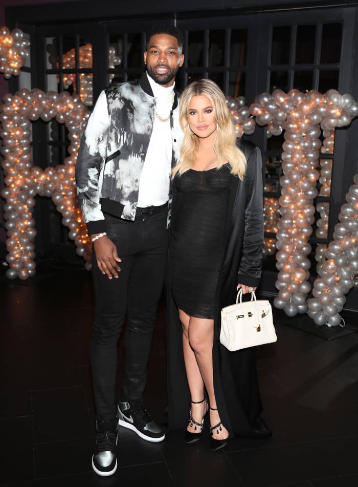 Tristan Thompson, in a black and white graphic vest with a white shirt and black pants, stands next to Khloe Kardashian, in a black dress