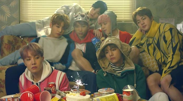 BTS sit huddled together in front of a table with party debris and a cake in the Spring Day music video