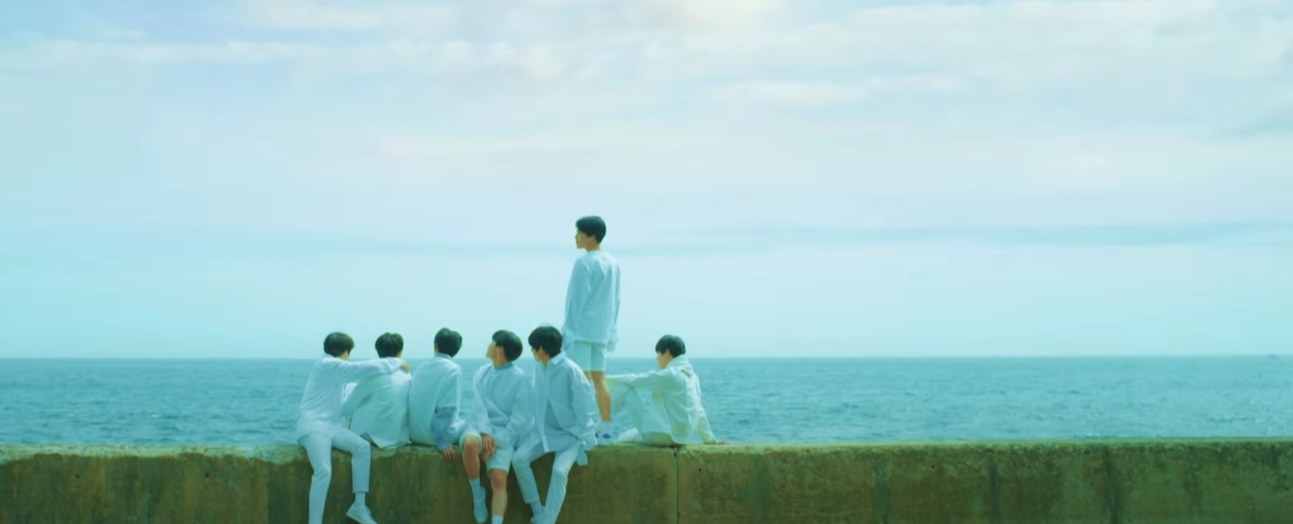 BTS all wear white and sit on a concrete wall by the ocean in the Euphoria music video