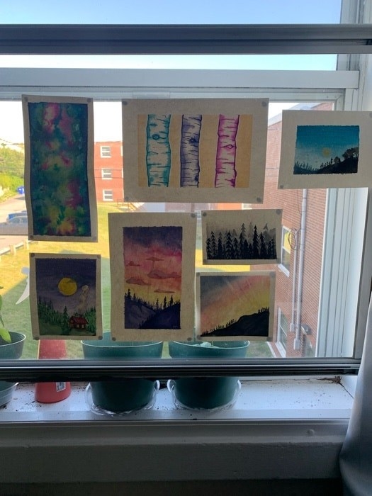 Seven watercolor paintings on white paper are taped to a clear window