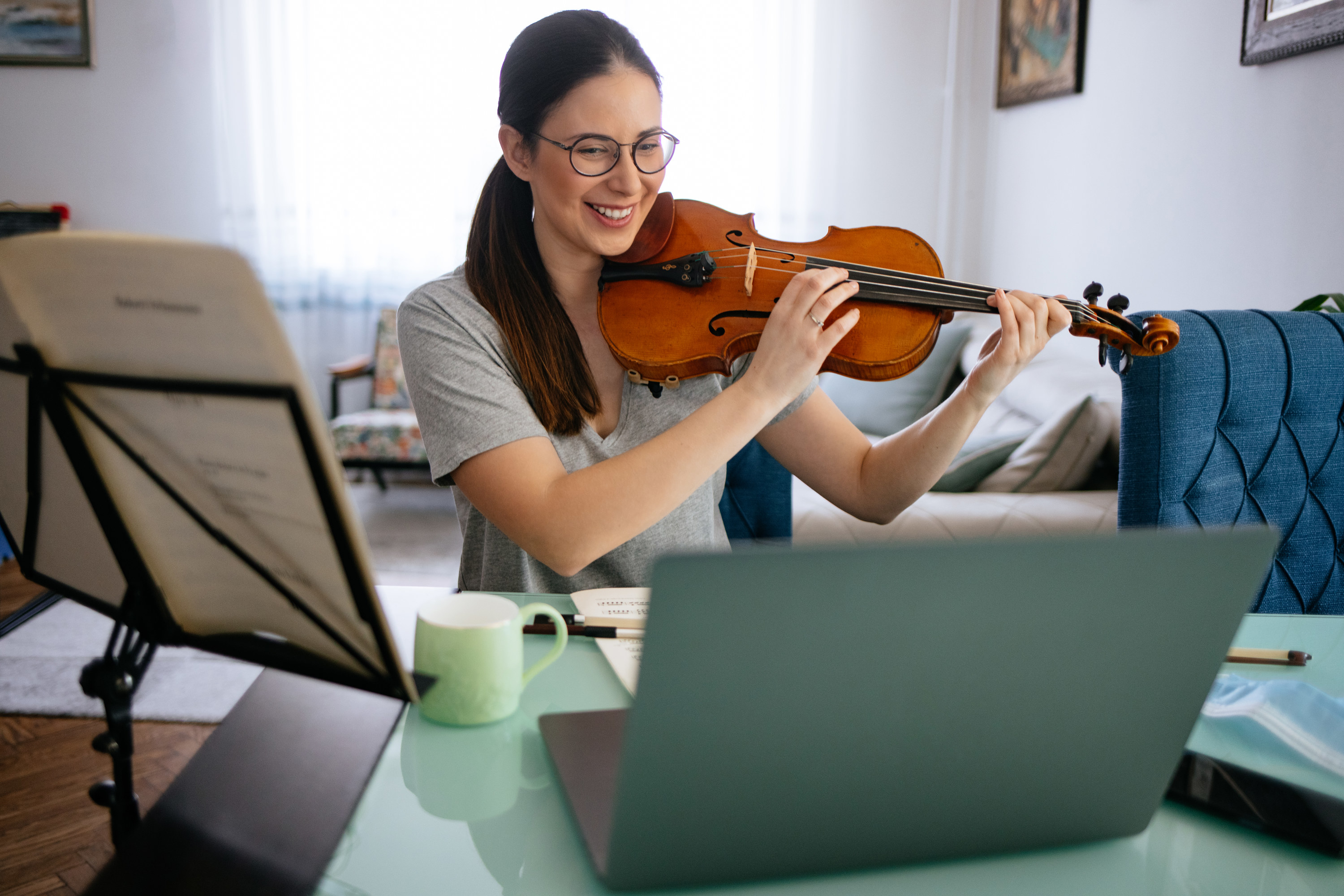 Woman smiles, while playing the violin in front of her laptop