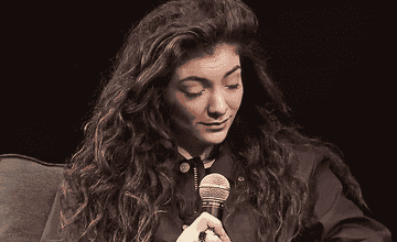 A seated Lorde holds a microphone while shrugging and looking to the side