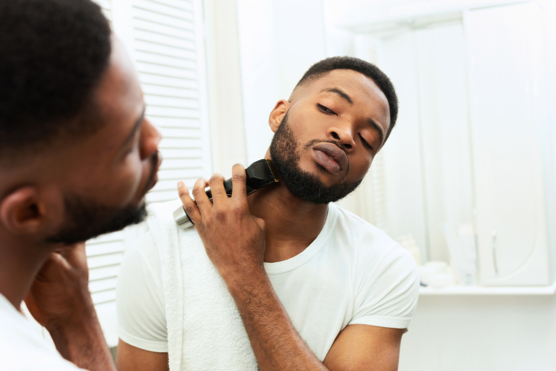 Young man looking at mirror and shaving beard with trimmer or electric shaver at bathroom