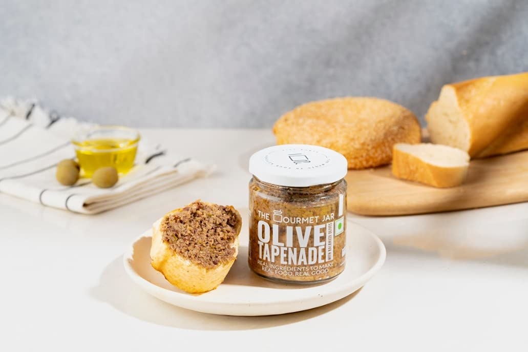 The jar of olive tapenade next to a slice of bread with tapenade spread on it.