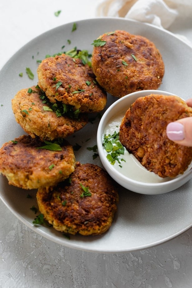 A hand dips a palm-sized chickpea fritter in tahini sauce; five other fritters surround it.