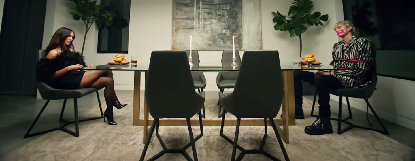 Megan, in a short black, fuzzy outfit and high heels, and MGK in black pants and white-and-black striped suit, sitting across from each other at a long table