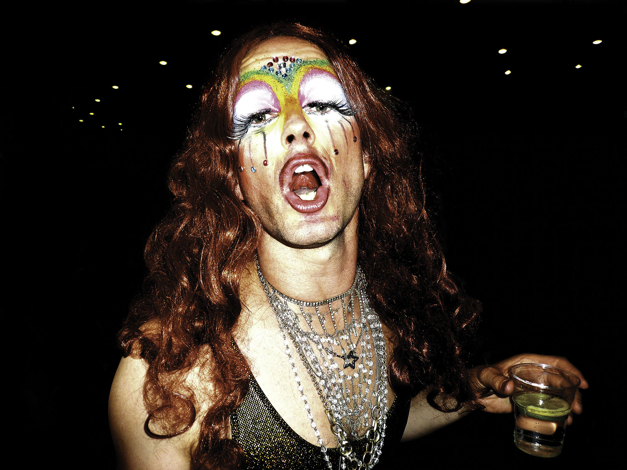 A man in drag holding a drink yells at the camera